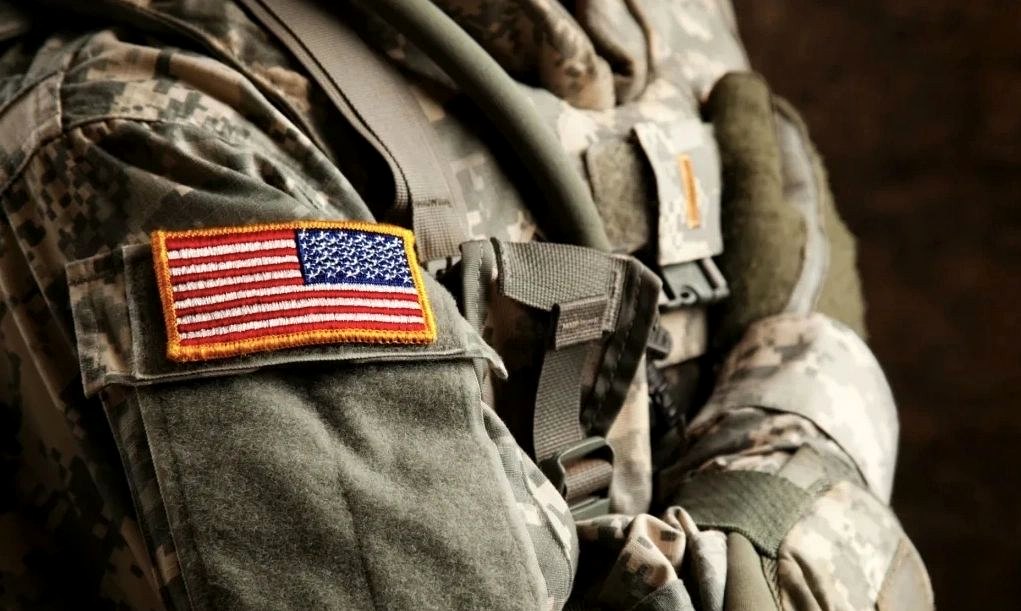 Shoulder of a service member in field uniform with a United States flag patch and load-bearing equipment.